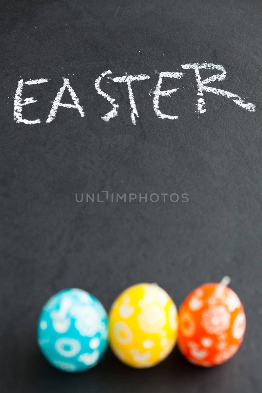 Closeup of three colorful Easter egg shaped candles and text written in chalk on dark background