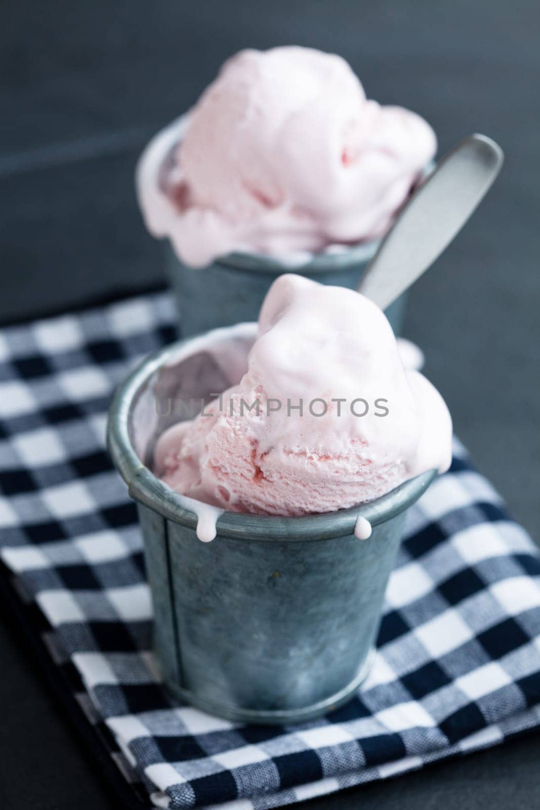 Closeup of strawberry ice cream in two metal cups on checkered serviette over dark background