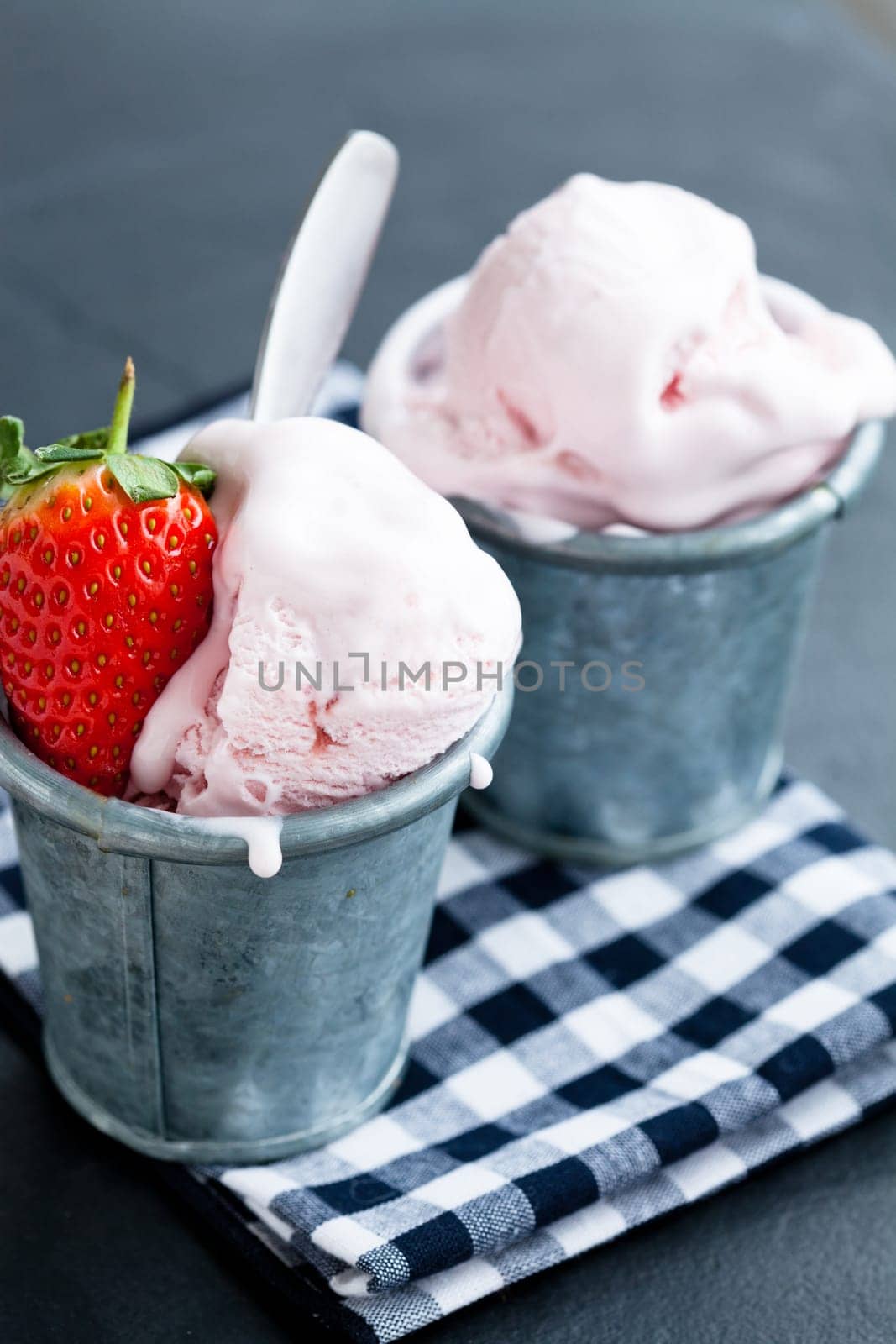 Closeup of strawberry ice cream and strawberry in two metal cups on checkered serviette over dark background