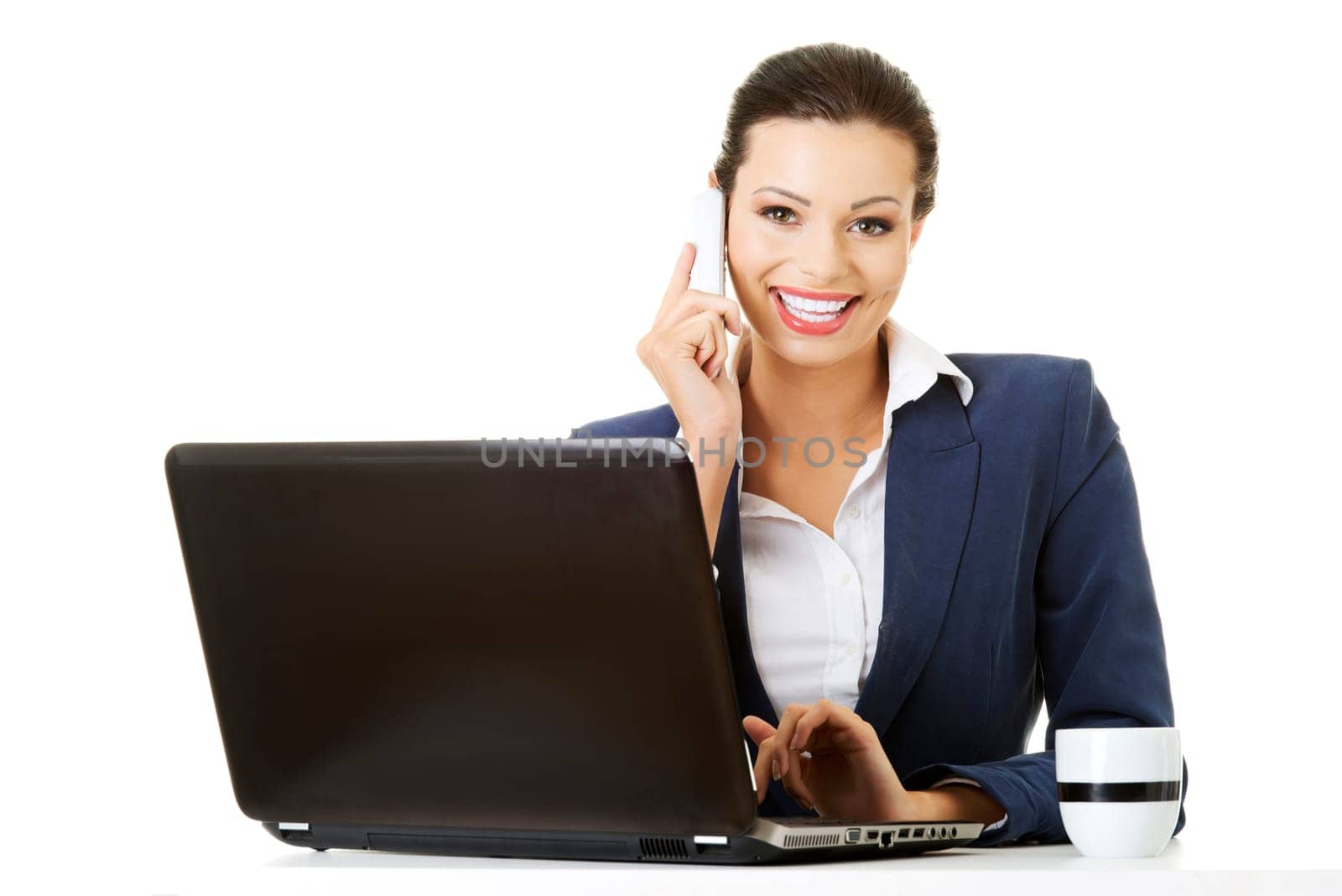 Smiling young business woman speaking on phone while using laptop, isolated on white