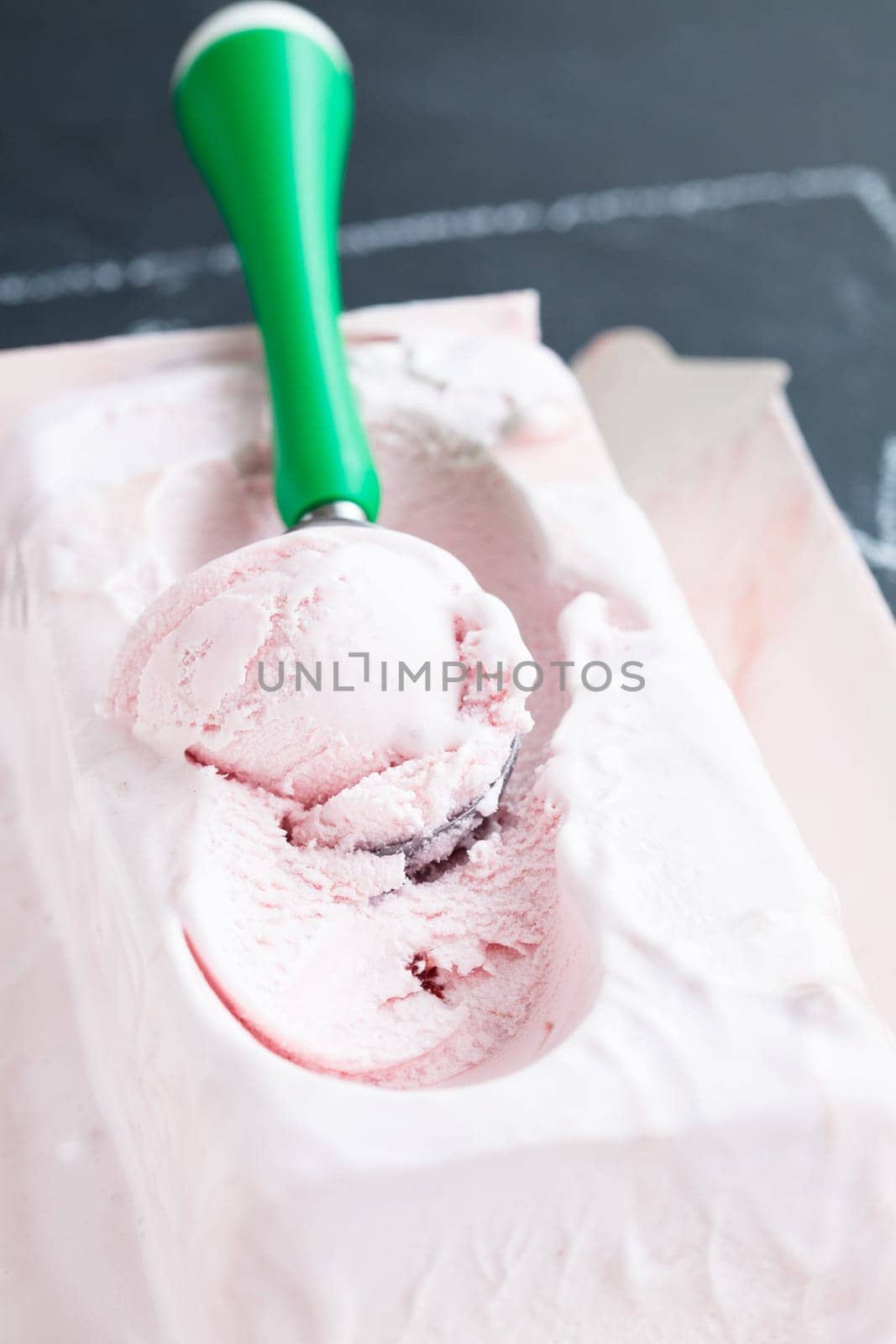 Closeup of tub of strawberry ice cream and scoop on dark background