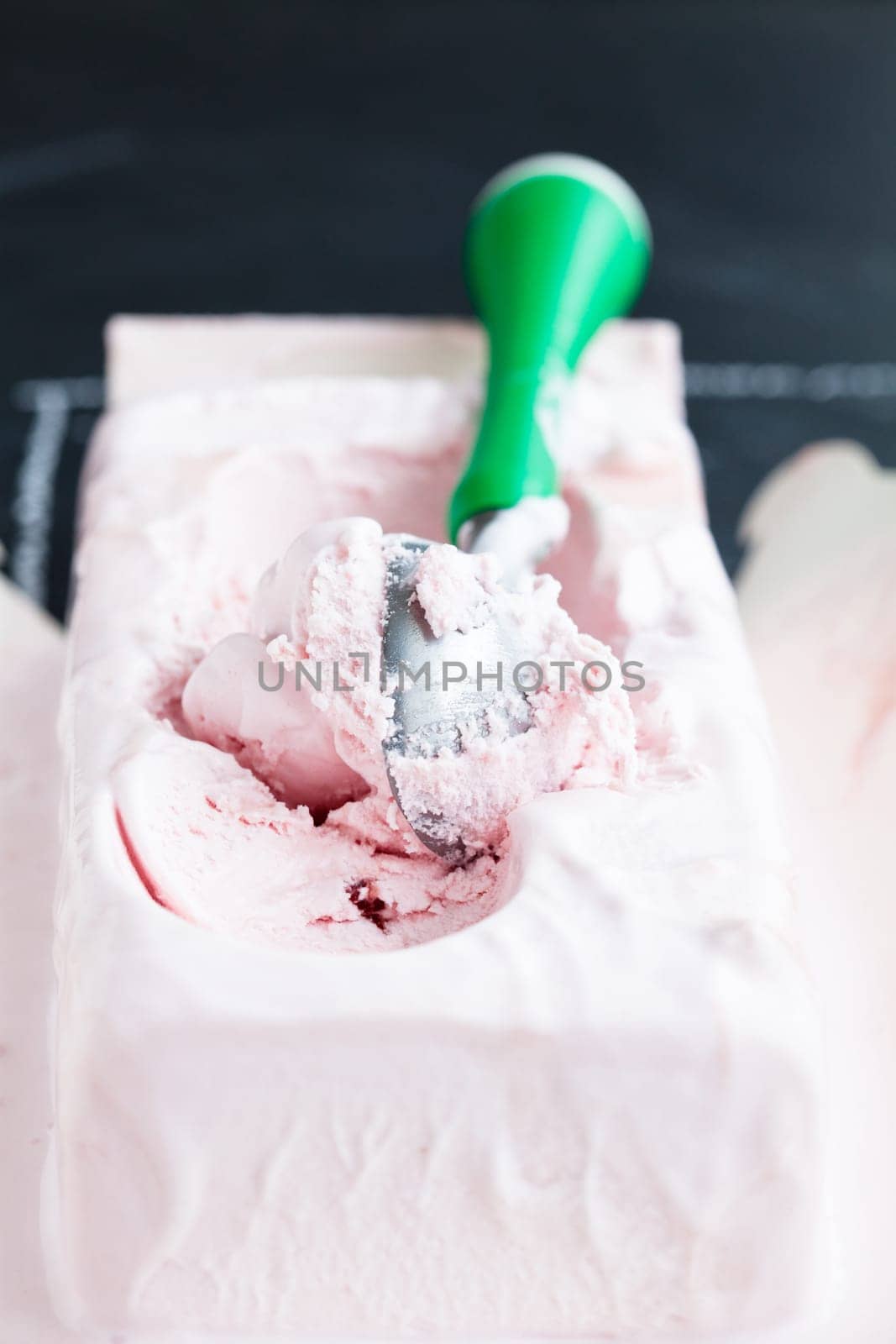 Closeup of tub of strawberry ice cream and scoop on dark background