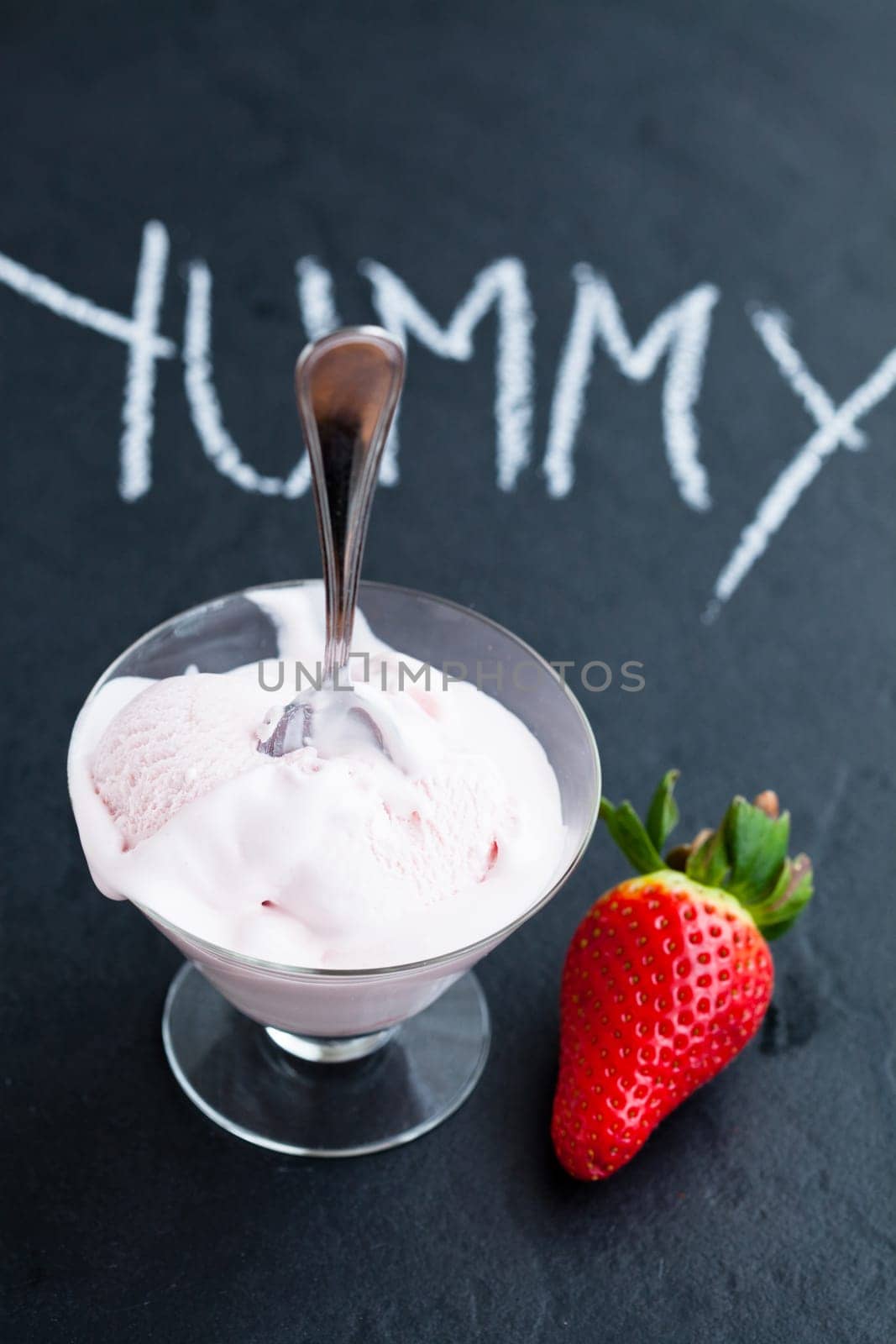Strawberry ice cream in glass bowl and whole strawberry on dark background with text written in chalk
