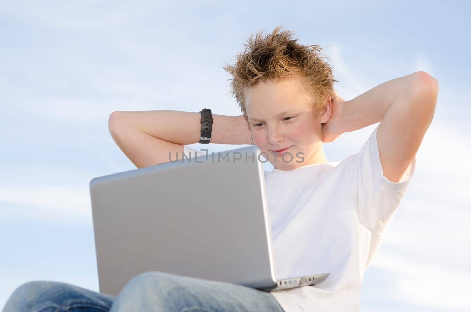 Fair-haired schoolkid leaning back with laptop against sky