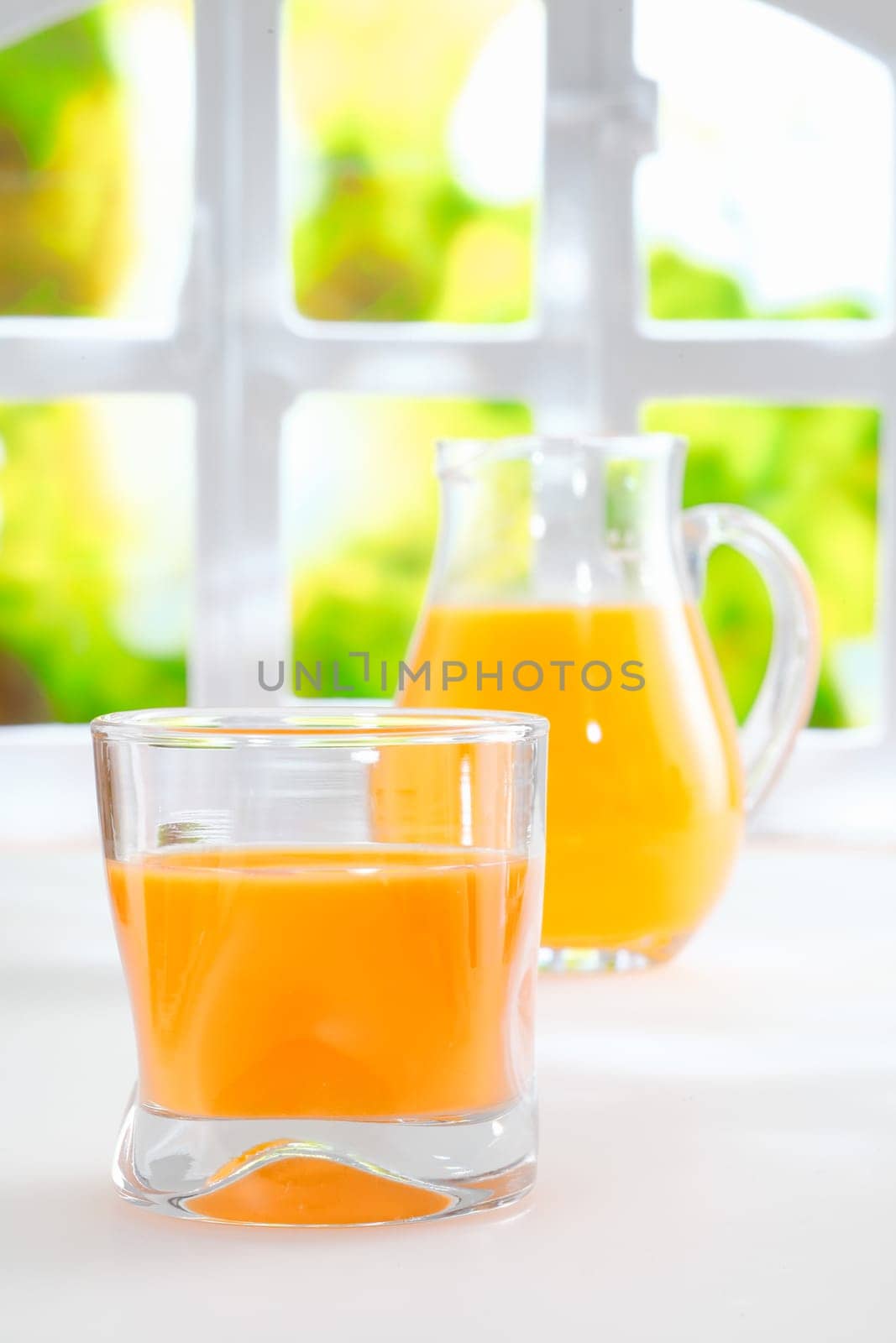 Healthy freshly squeezed orange juice in a glass tumbler with a glass jug full of juice standing in front of a fresh summery window