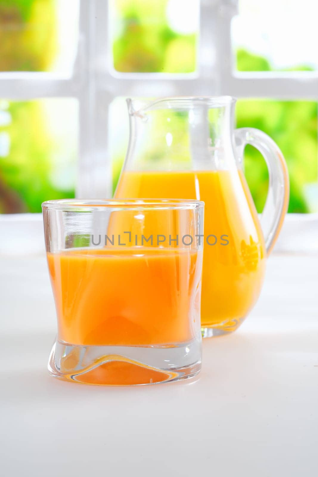 Tumbler of fresh healthy orange juice standing on a kitchen table alongside a jug full of juice with a sunny window and greenery behind