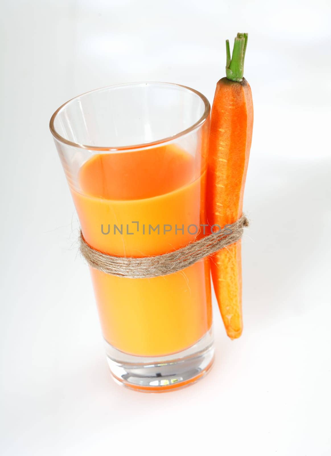 Tall glass of fresh vegetarian carrot juice with an attached whole carrot standing on a white studio background