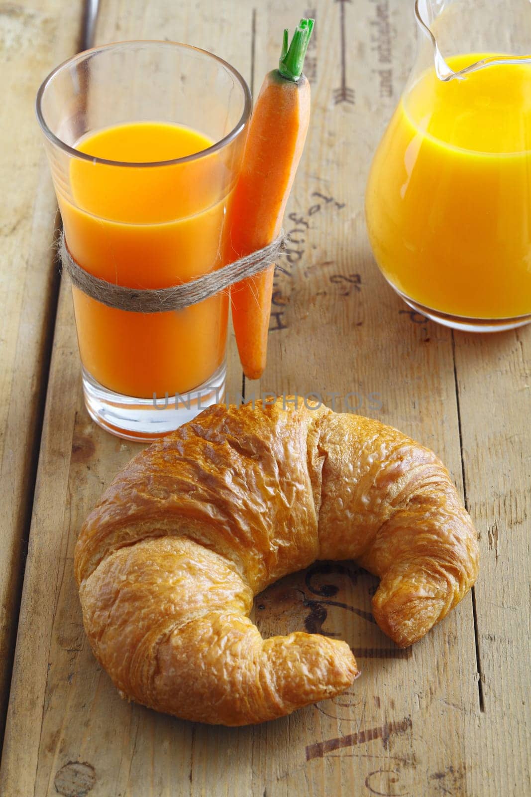 Crisp flaky golden croissant lying on an old textured wooden tabletop with a tall glass of carrot juice for a healthy snack