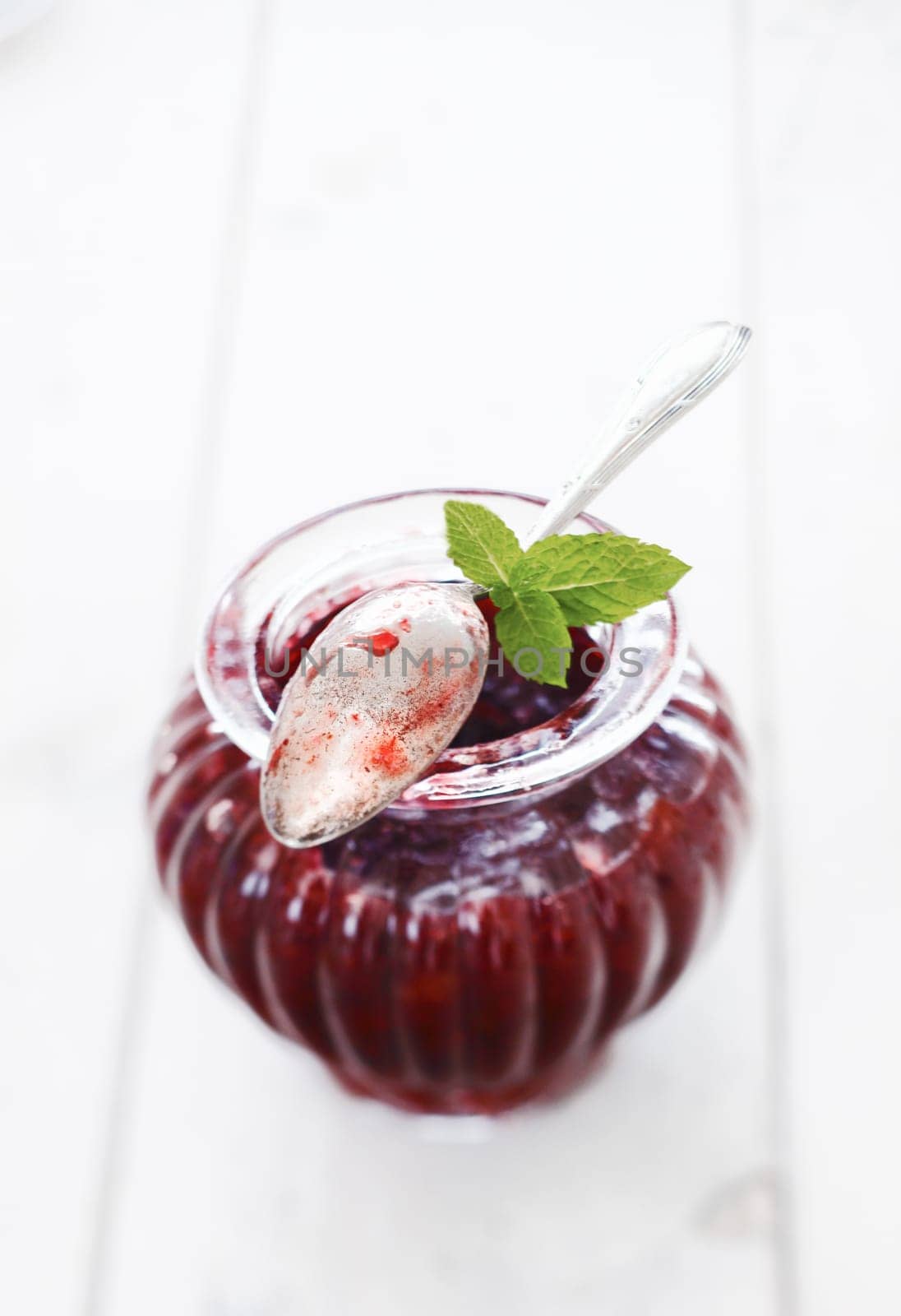 Elegant fluted glass jar of berry jam with a used silver teaspoon coated in jam resting on top of the rim with three fresh mint leaves