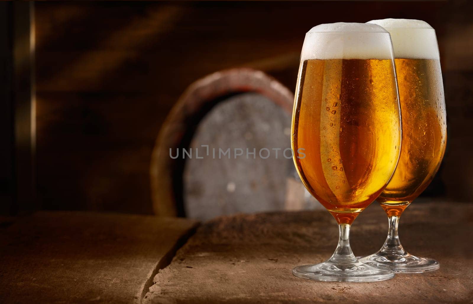 Two glasses of fresh foamy beer on a table in a vintage beer cellar with a barrel in the background
