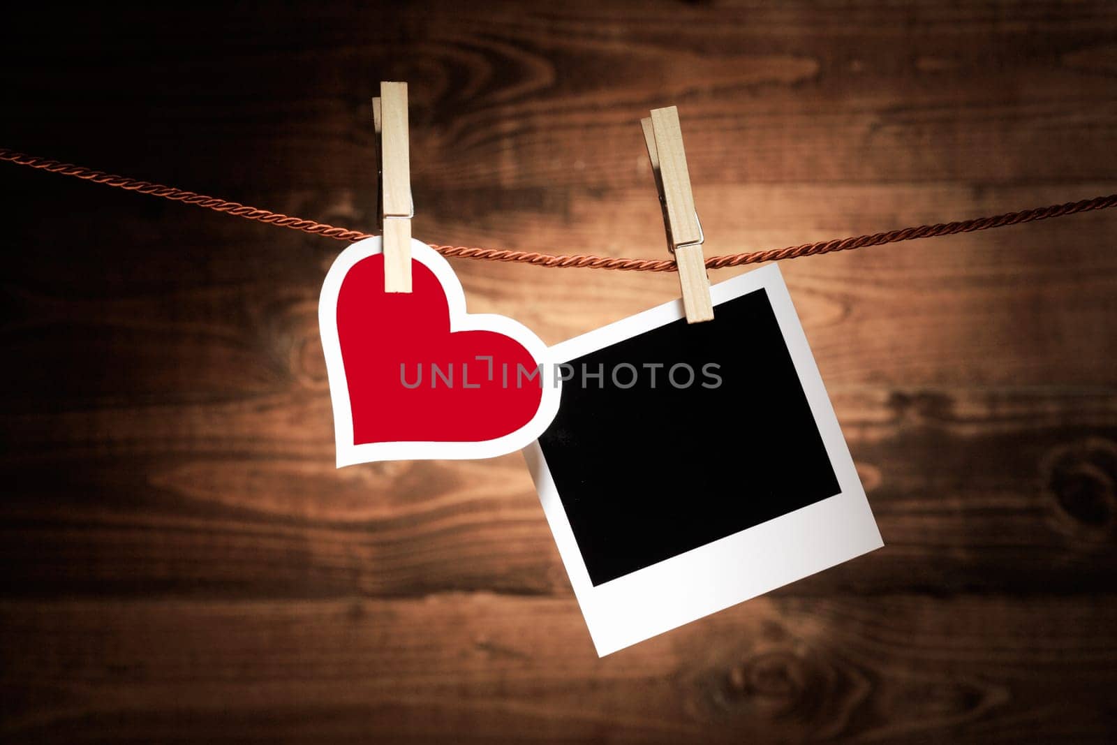 A red and white heart hangs from a wooden clothespeg alongside a blank instant photo frame on a rustic wooden background for your nostalgic romantic memories or Valentines message