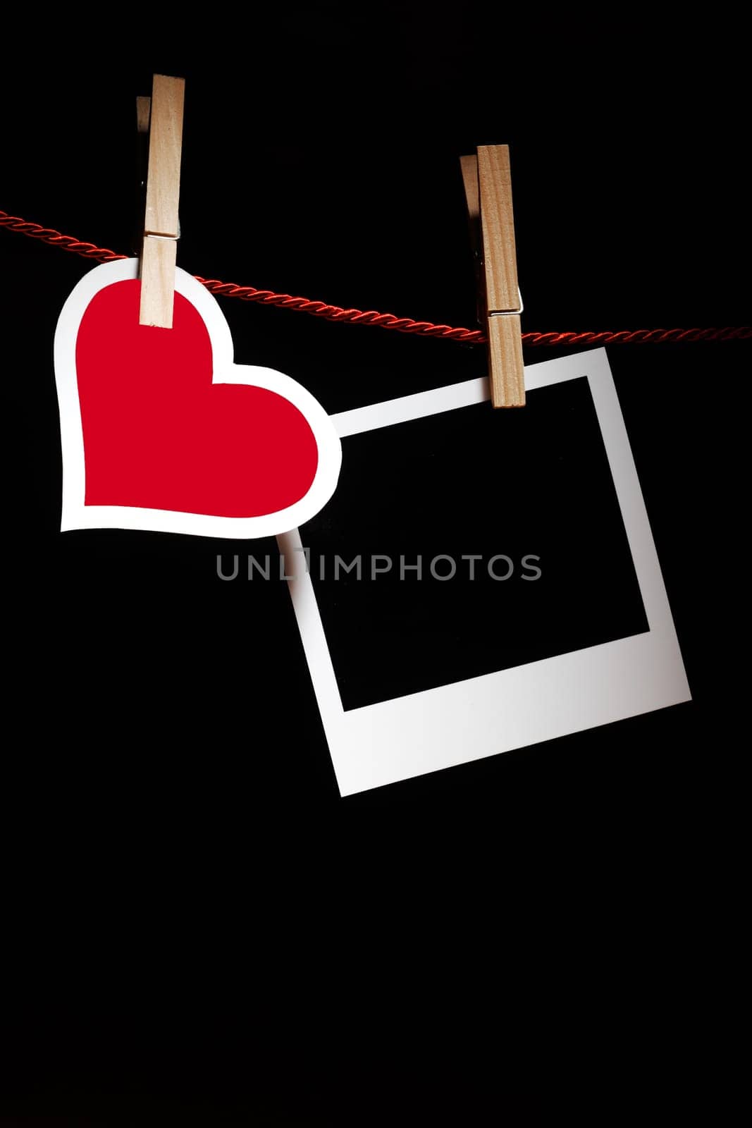 Pretty red and white heart hanging with a blank instant photo frame from wooden pegs on a line against a dark background with copyspace for your Valentine or romantic message