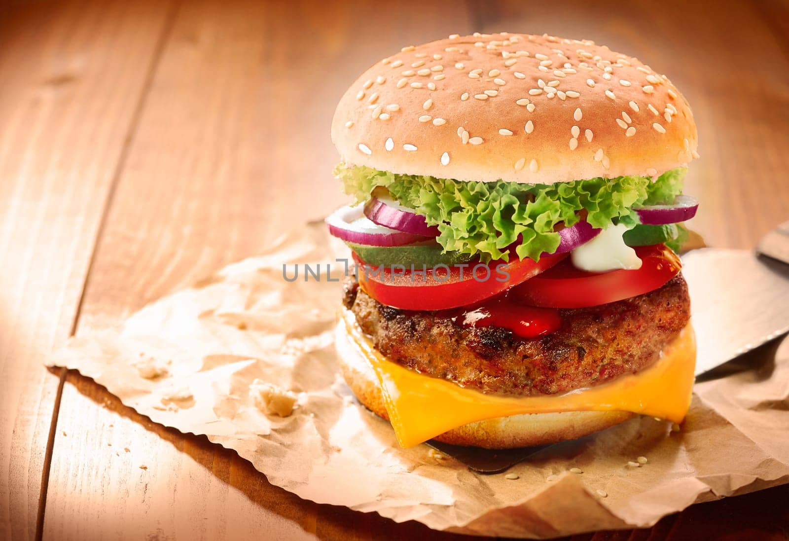 Delicious cheeseburger stacked high with a juicy beef patty, cheese, fresh lettuce, onion and tomato on a fresh bun with sesame seed standing on brown paper on a wooden tabletop with copyspace