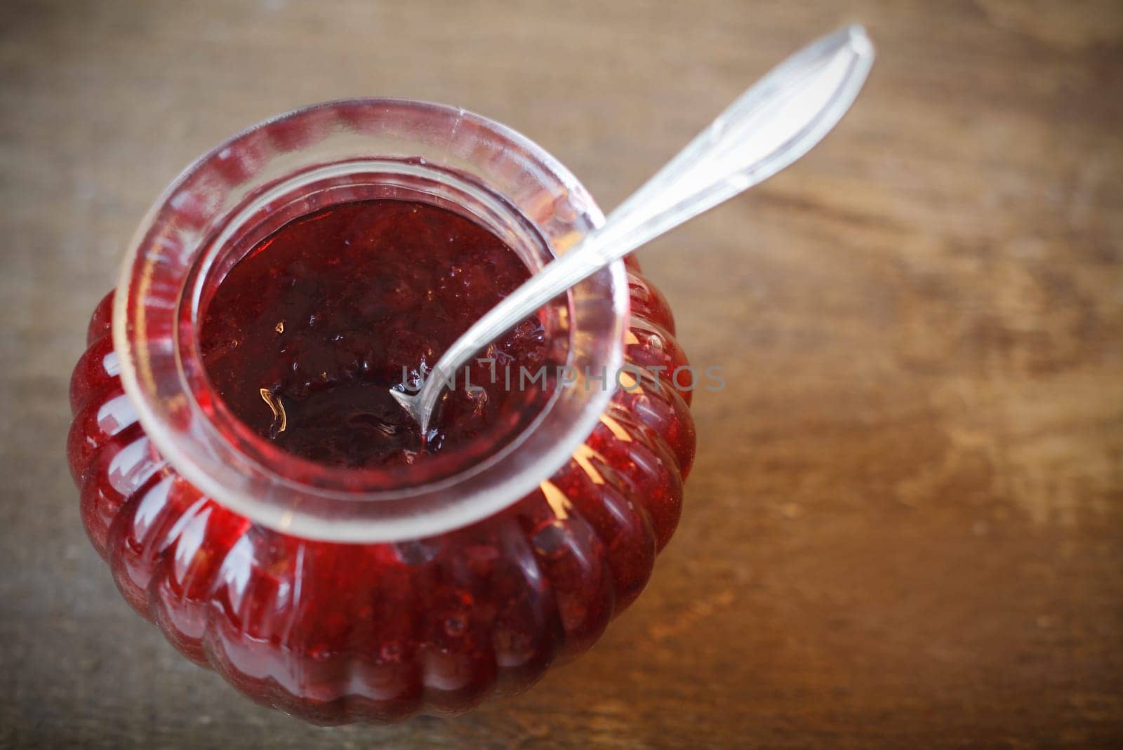 Overhead view of a decorative fluted glass jar filled with fresh fruity strawberry jam and a silver serving spoon on a textured wooden surface with copyspace