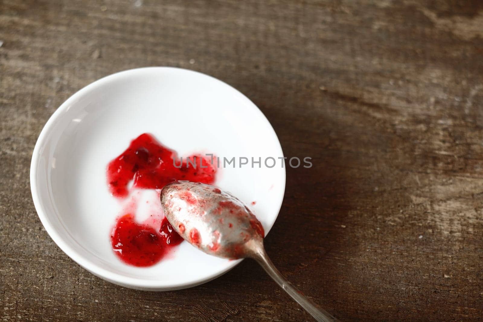 Silver teaspoon resting on a small white dish with samples of fresh fruity strawberry jam taken while preparing a batch in the kitchen