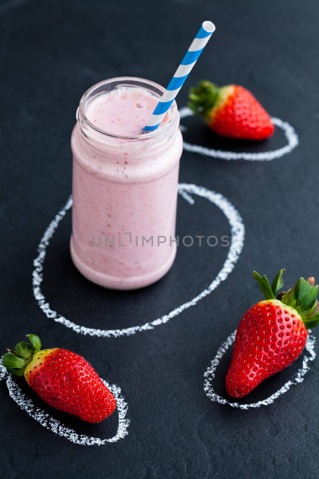 Strawberry smoothie with striped straw and whole strawberries on dark background