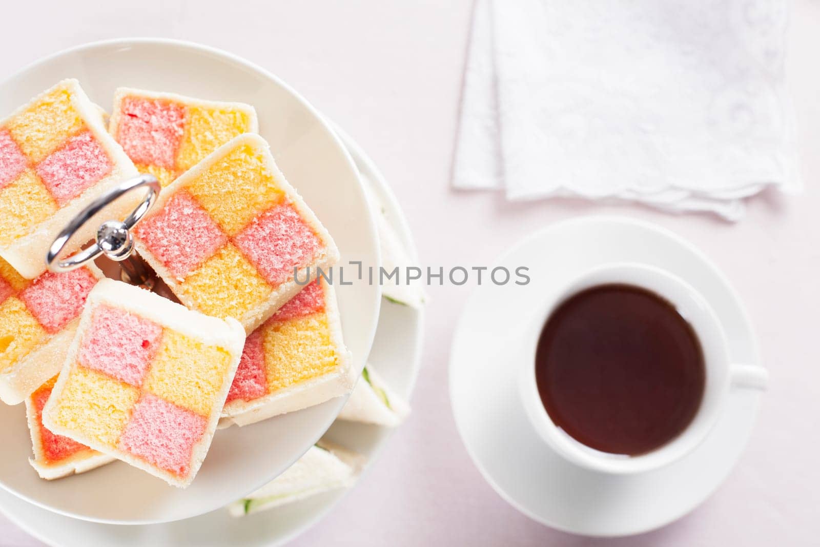 Battenberg cake on a two tiered serving plate and a cup of tea on the table