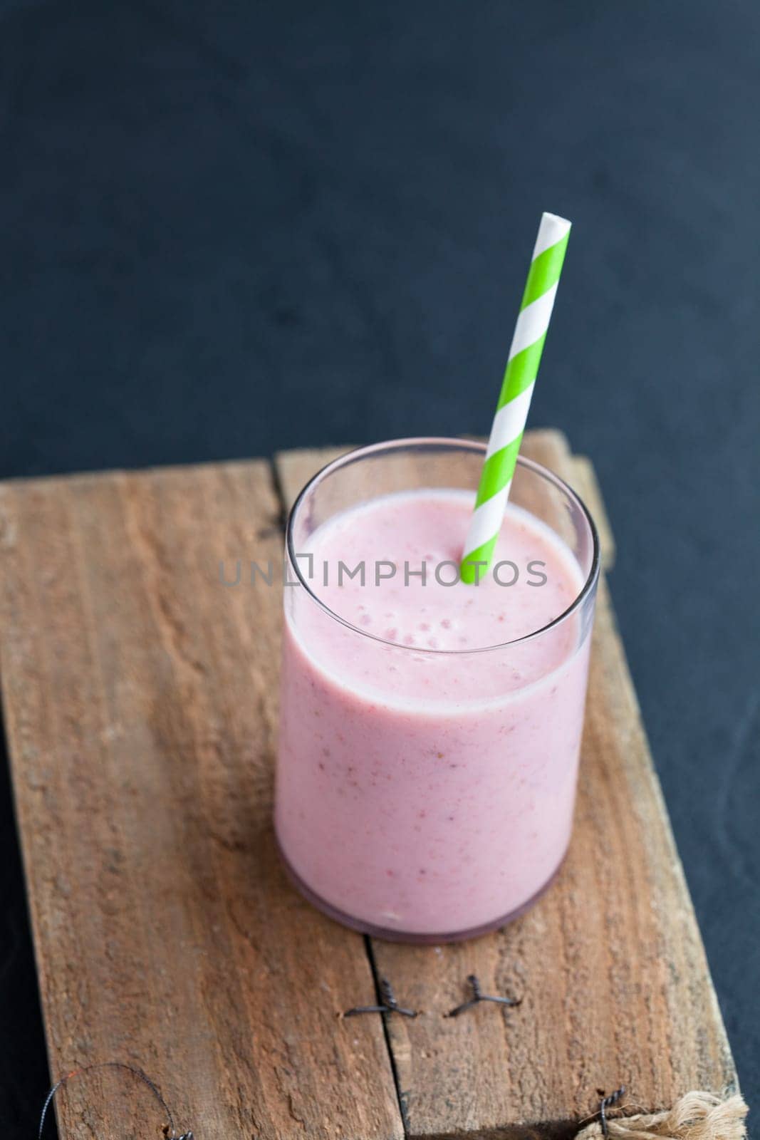 Strawberry smoothie with striped straw on wooden serving tray on dark background