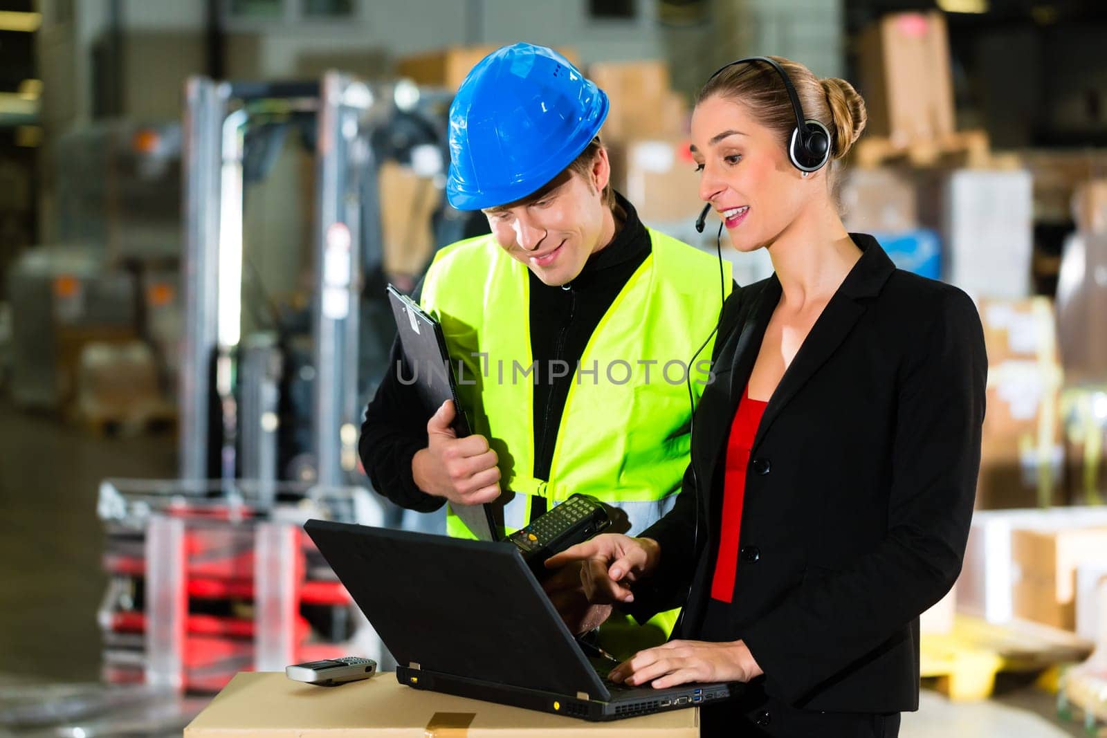 Teamwork - warehouse clerk or warehouse worker and customer service representative using laptop and headset in a warehouse of a freight forwarding company,she points at the camera