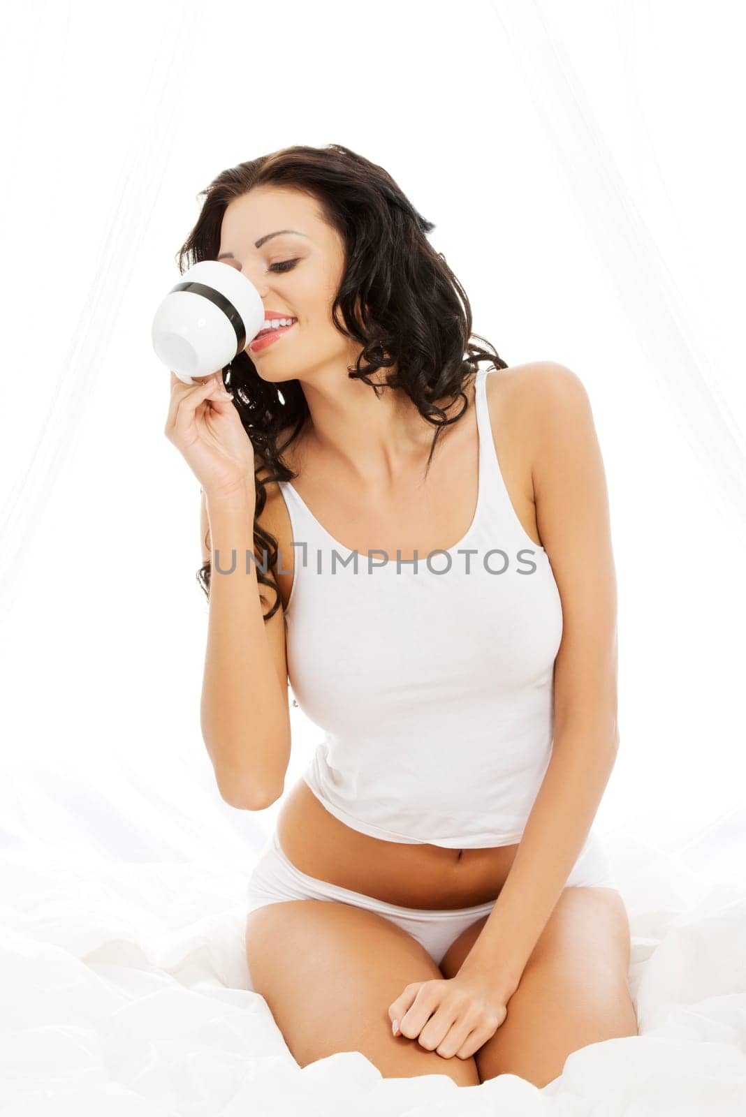 Beautiful young woman sitting on bed with big mug of something hot