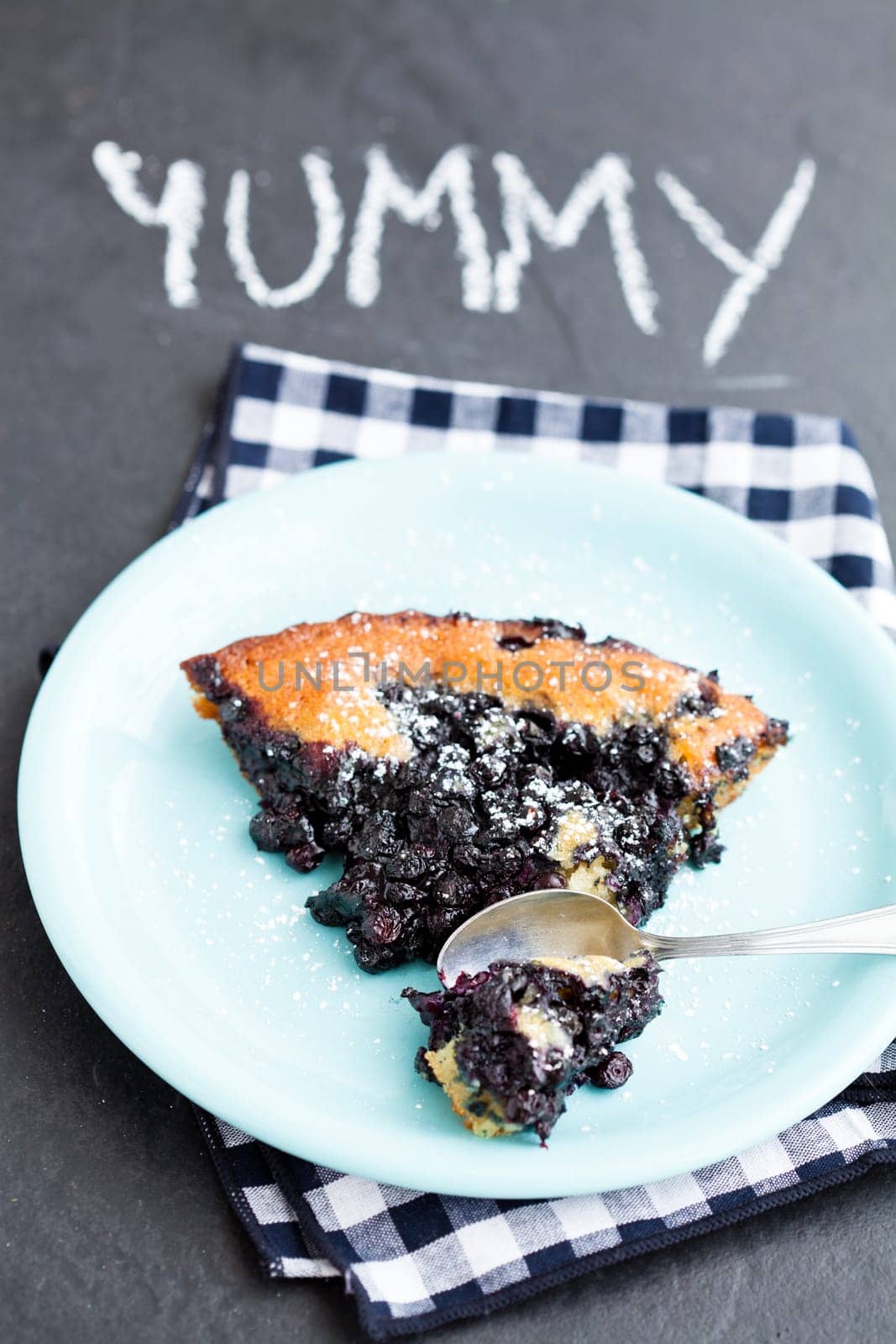 Closeup of slice of delicious blueberry pie on plate with spoon on dark surface with word yummy written in chalk