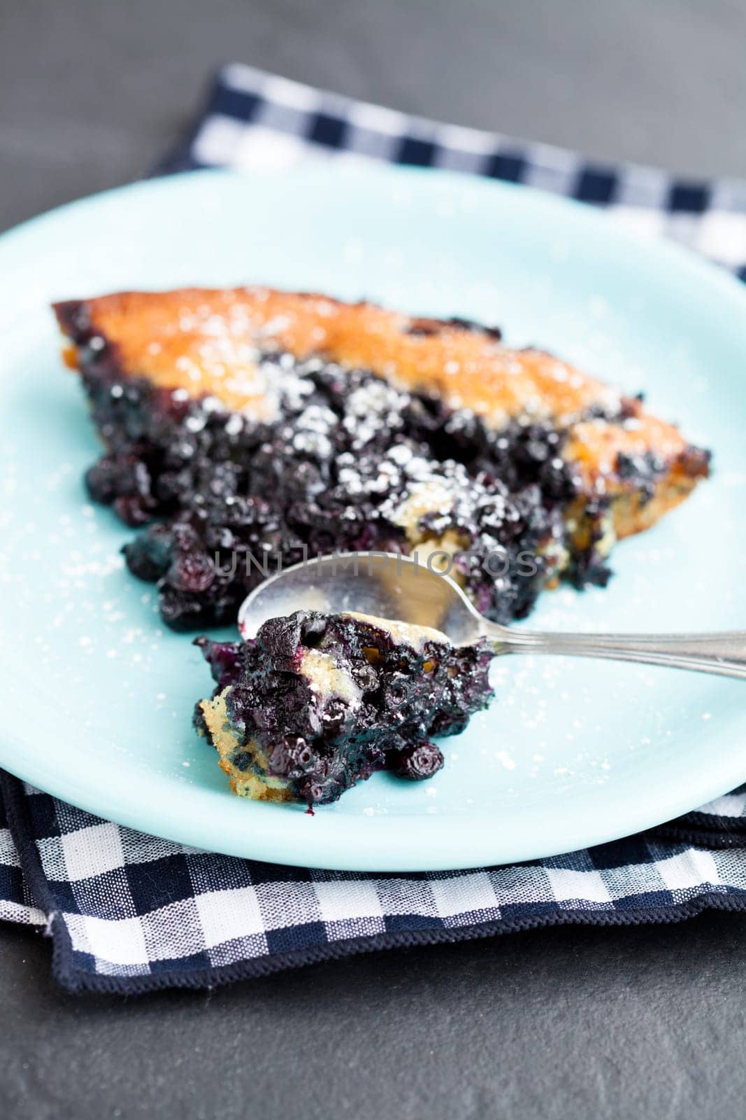 Closeup of slice of delicious blueberry pie on plate with spoon resting on checkered napkin