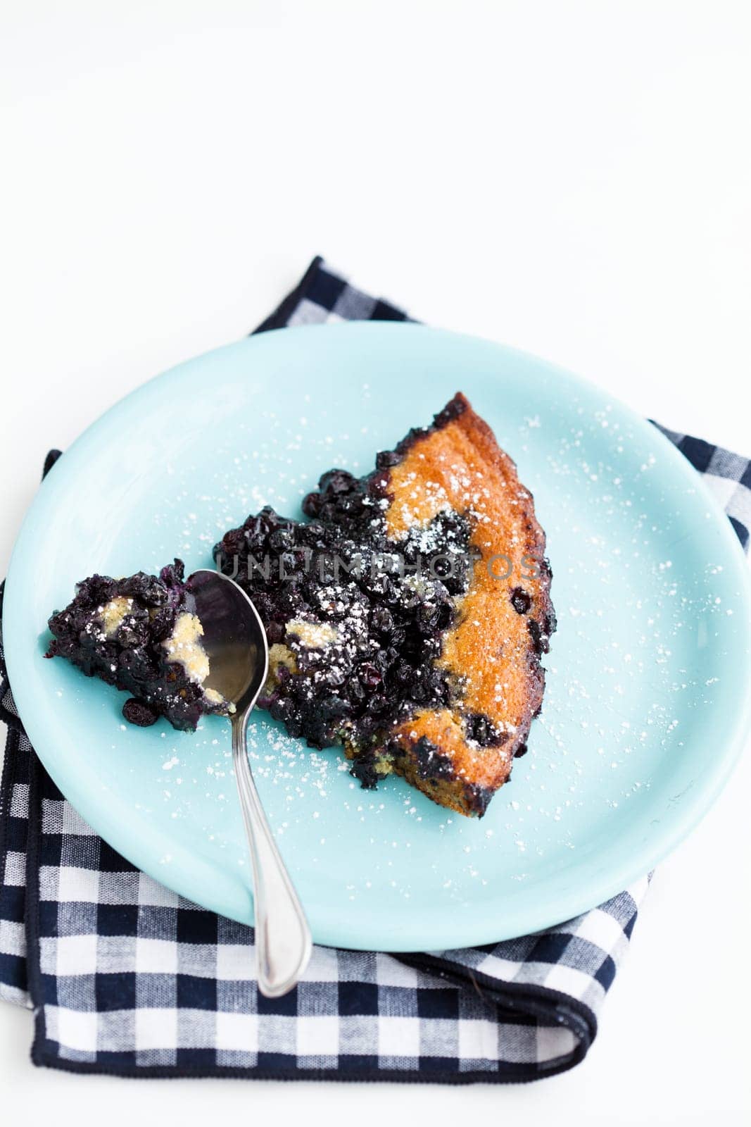 Closeup of slice of delicious blueberry pie on plate with spoon resting on checkered napkin over light background