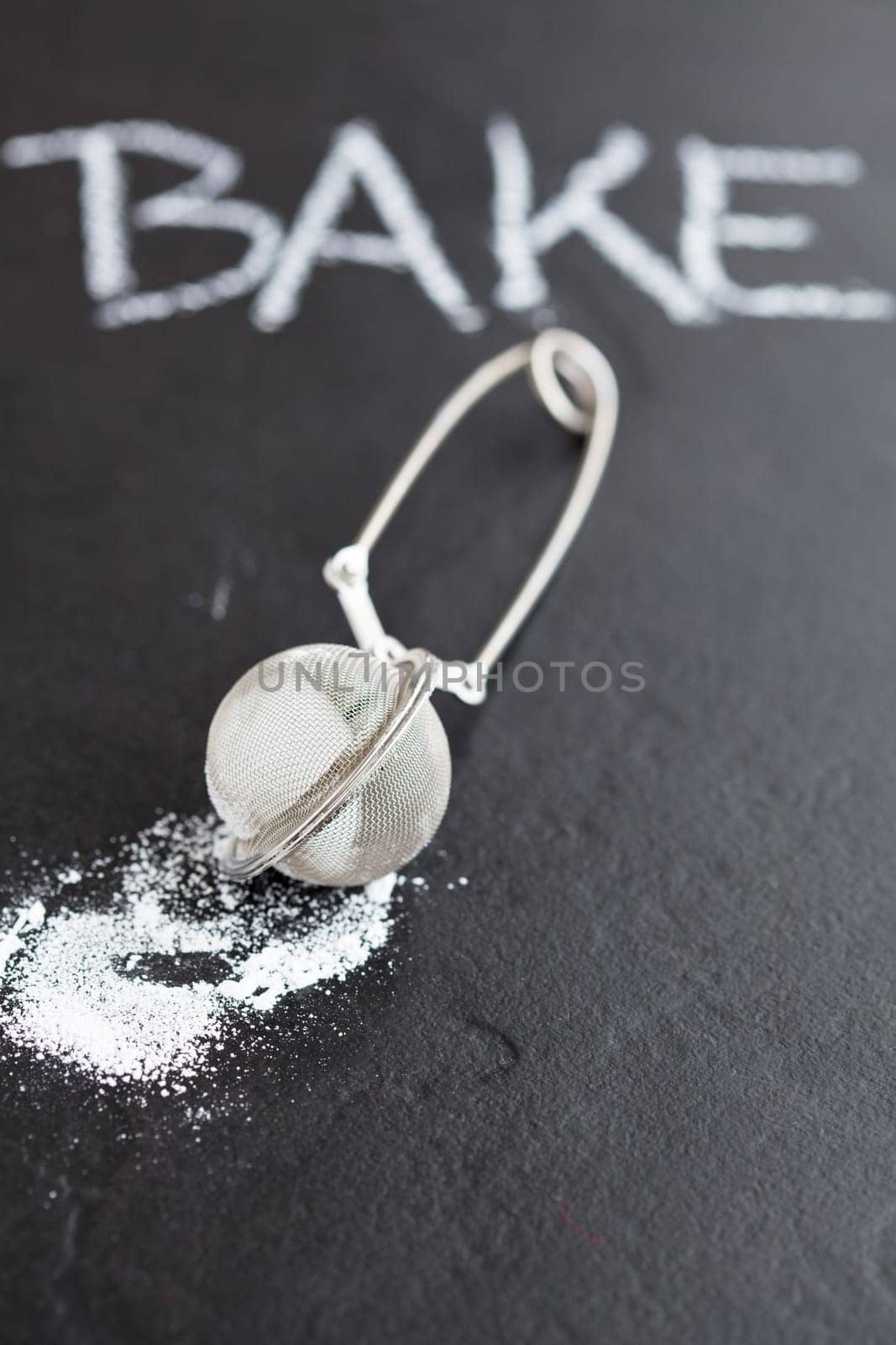 Closeup of sieve and icing sugar with word bake written in chalk on dark textured background