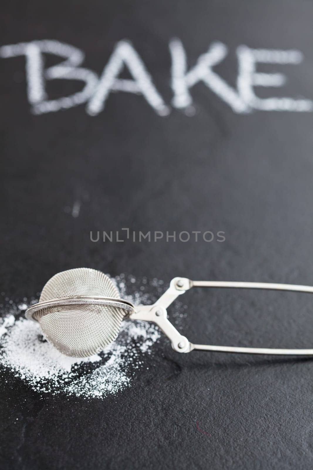 Closeup of sieve and icing sugar with word bake written in chalk on dark textured background