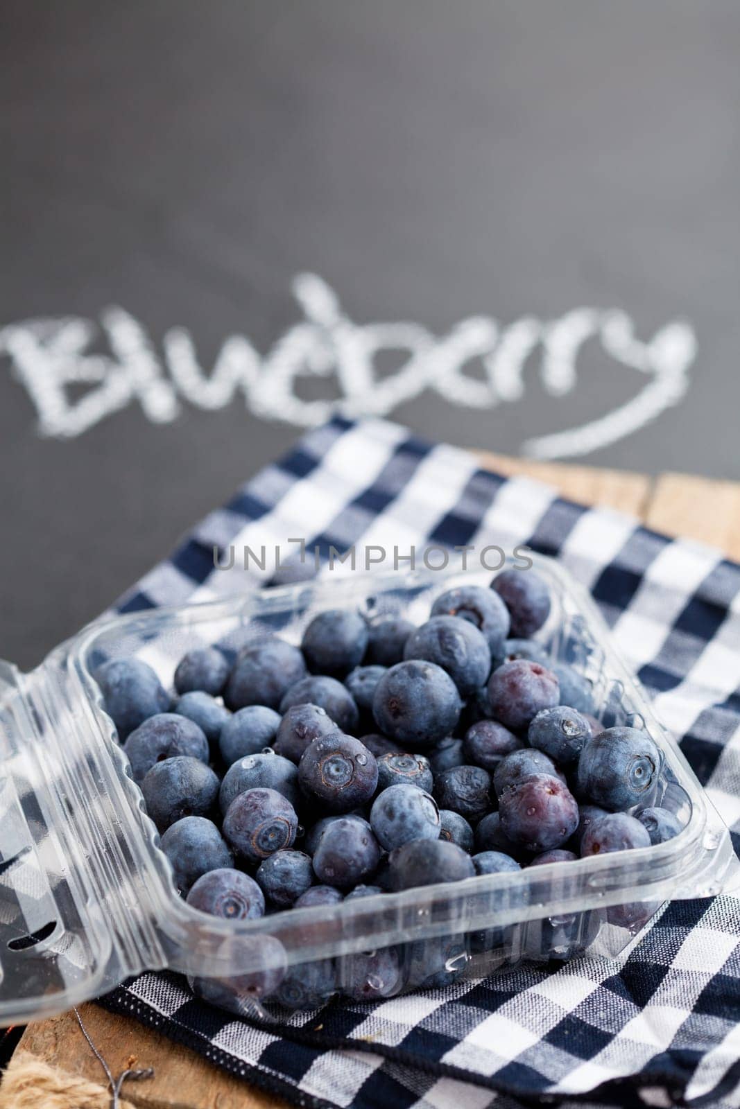 Closeup of fresh blueberries in plastic container on checkered napkin with word blueberry written in chalk on dark surface