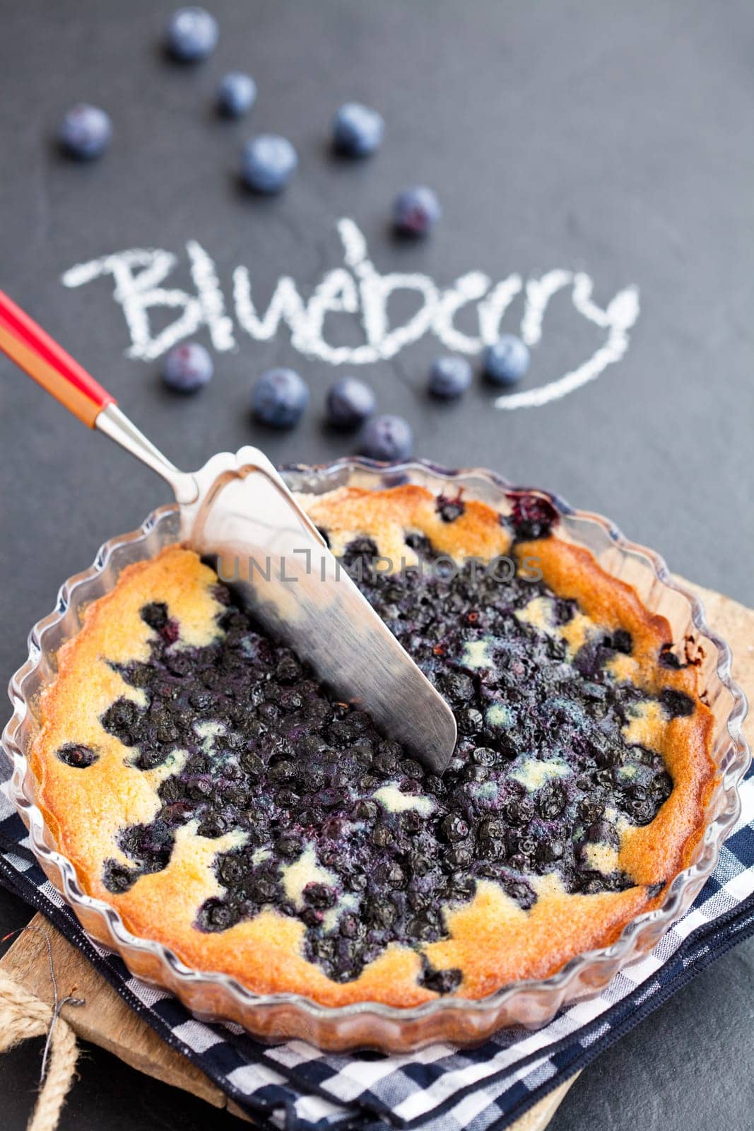 Blueberry pie in glass dish and server with word blueberry written in chalk on dark background