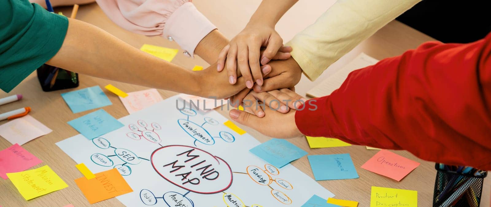 A group of business people putting their hands together at meeting room on table with mind map scatter around. Startup group showing unity teamwork and friendship. Close up. Focus on hand. Variegated.