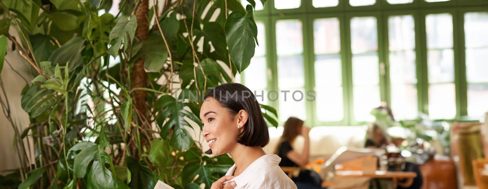 Vertical shot of asian brunette girl sitting elegant with book, reading in cafe and smiling, drinking coffee by Benzoix