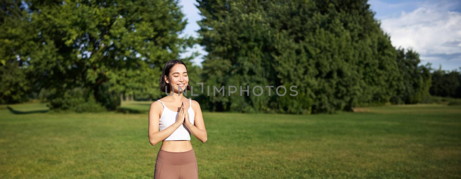 Vertical shot of smiling asian girl doing yoga, stretching exercises in park, standing on rubber mat on fresh air.