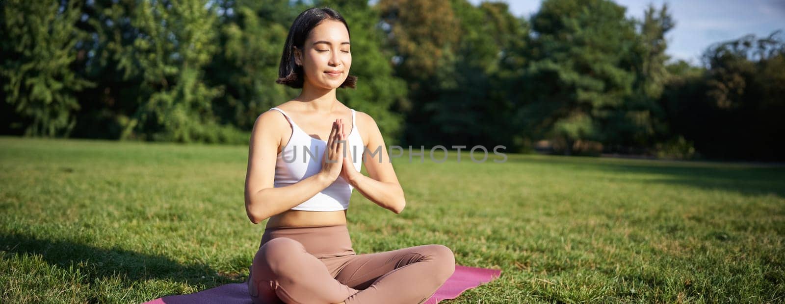 Woman meditating on lawn in park, sitting on sports mat, relaxing, breathing fresh air.