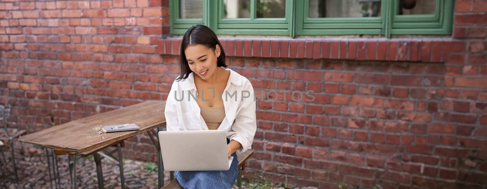 Vertical shot of asian woman working in outdoor cafe, sitting with laptop on bench and drinking coffee by Benzoix