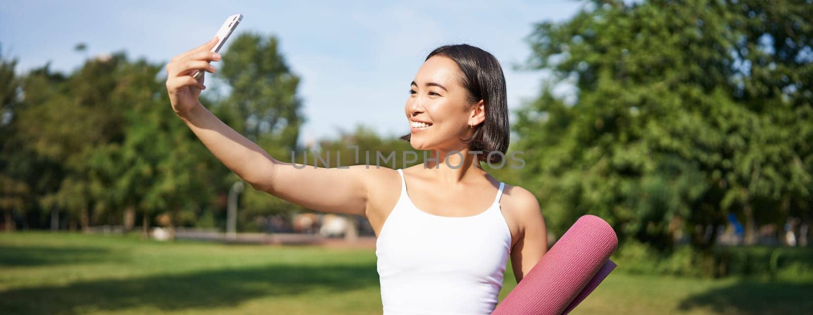 Sporty fitness girl takes selfie with rubber yoga mat in park, does workout and shares photos on social media.