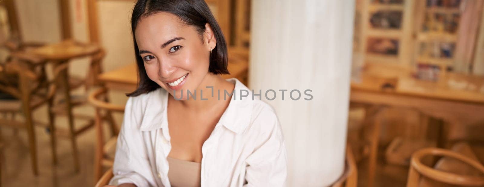 Vertical shot of young smiling woman with book, reading in cafe, enjoying morning breakfast coffee and croissant.