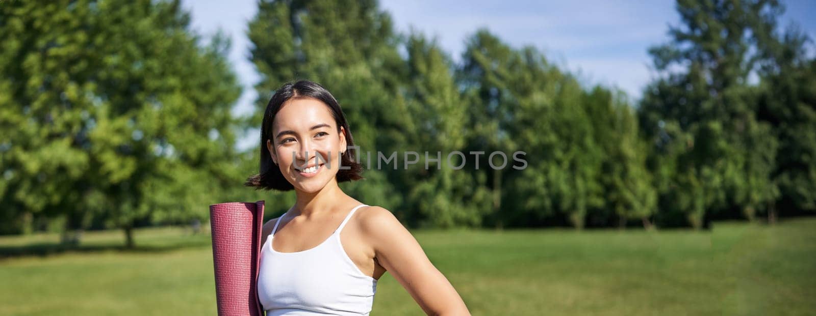 Vertical portrait of young asian fitness girl walks with rubber mat for yoga, goes on training session on fresh air in park, wears sports clothing.
