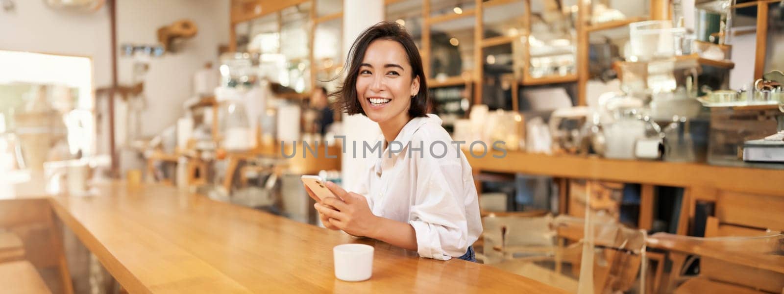 Portrait of young brunette woman, sitting with coffee and using smartphone in a cafe, chatting on mobile phone by Benzoix