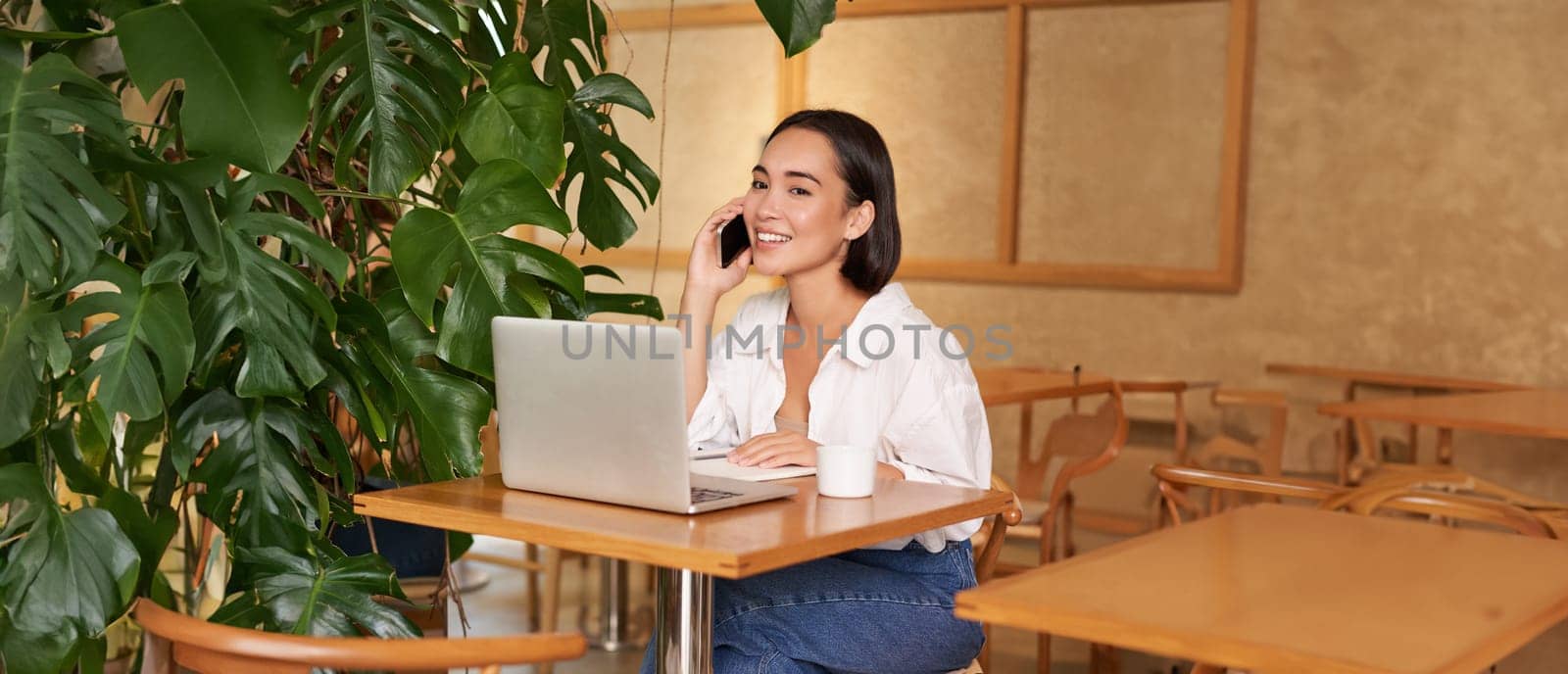Young female cafe manager, owner sitting with laptop, answering phone calls and smiling friendly by Benzoix