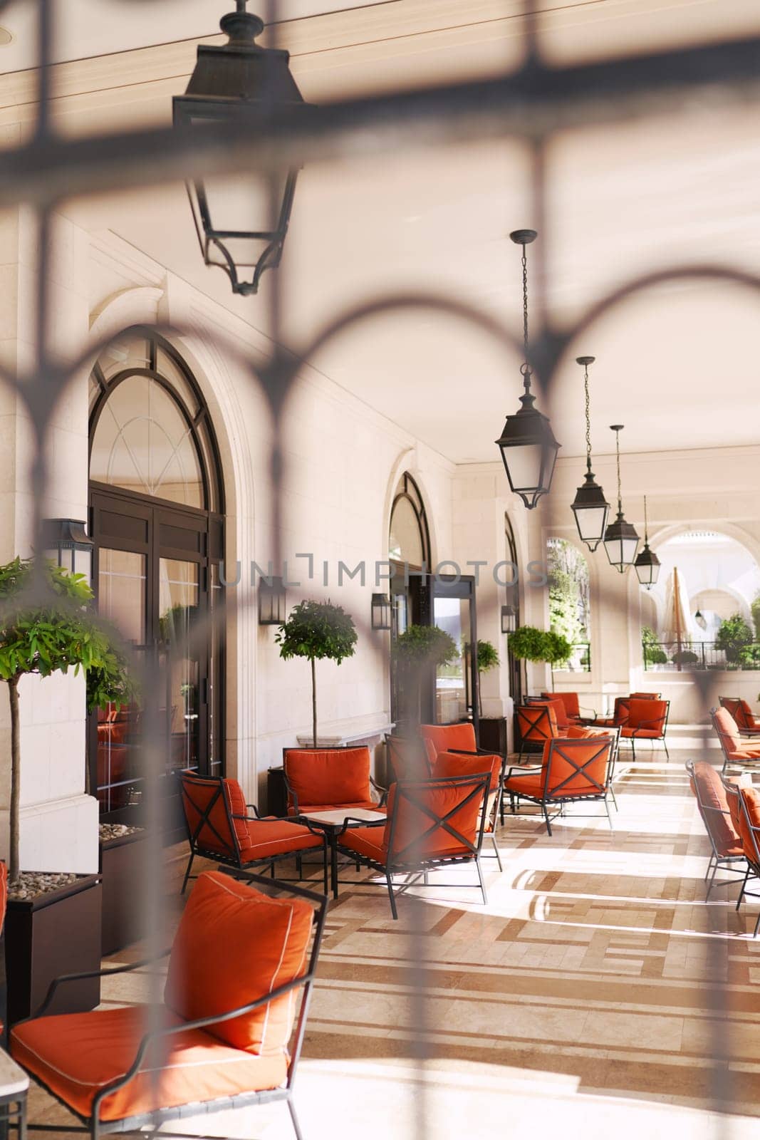 View through the wrought-iron lattice onto the hotel terrace with soft chairs and tables by Nadtochiy