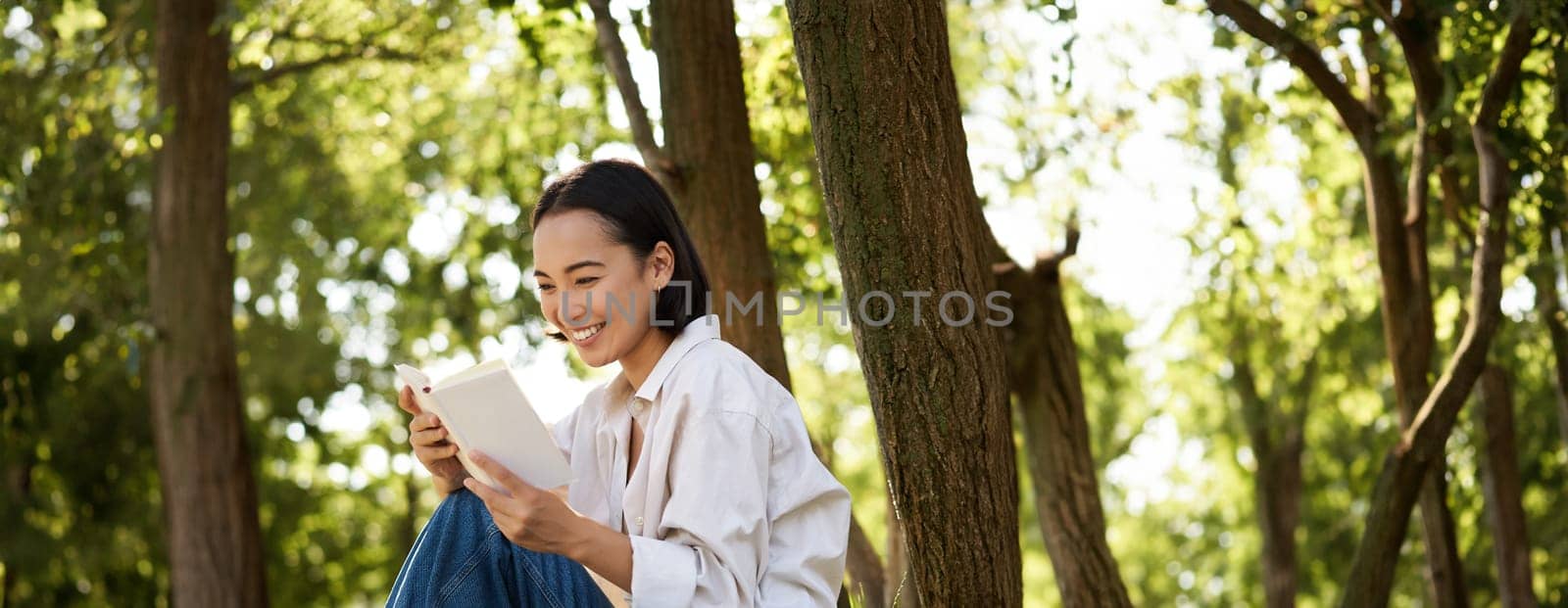 Vertical shot of happy asian woman relaxing outdoors in park, reading her book and sitting under tree shade on warm sunny day.