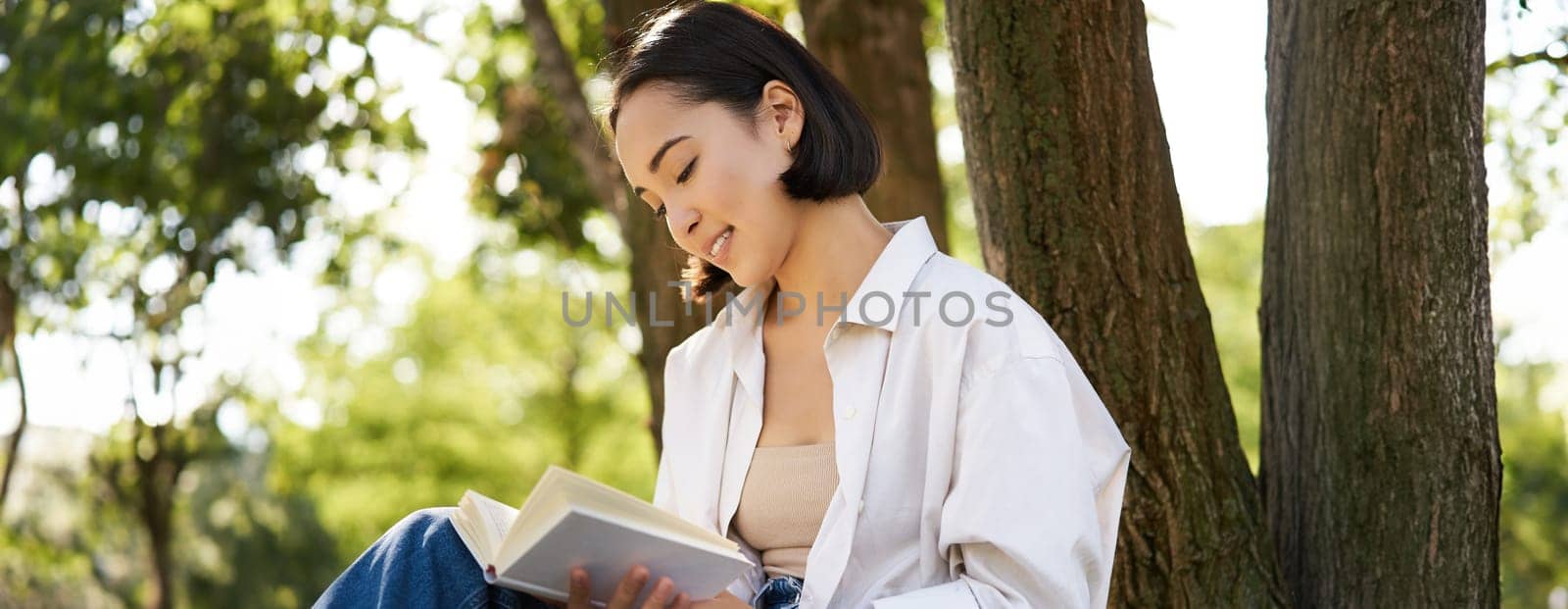 Beautiful smiling girl reading book in park on sunny day, sitting near tree on lawn and relaxing outdoors.