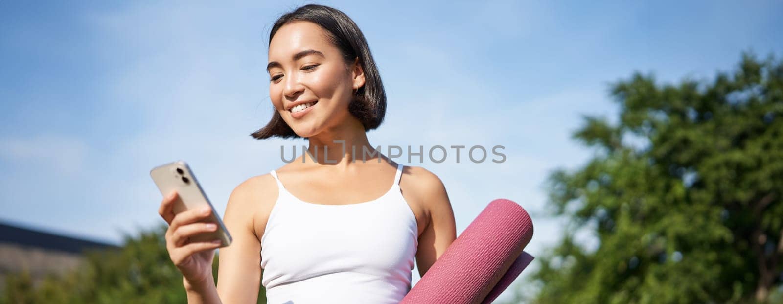 Portrait of smiling asian woman with yoga mat, looking at her smartphone and reading on application, standing in park wearing sport uniform.