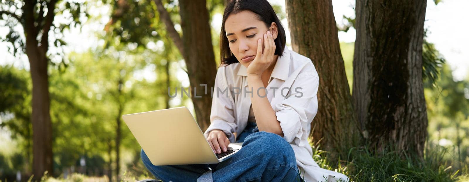 Portrait of asian woman sitting on grass near tree, using laptop, working, doing homework remotely on sunny summer day.