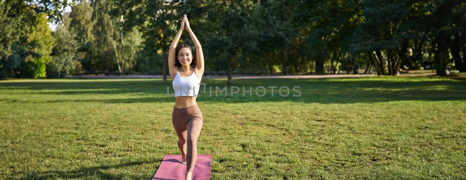 Vertical shot of asian woman standing in tree pose, making yoga asana on fresh air, wellbeing training in park on rubber mat.