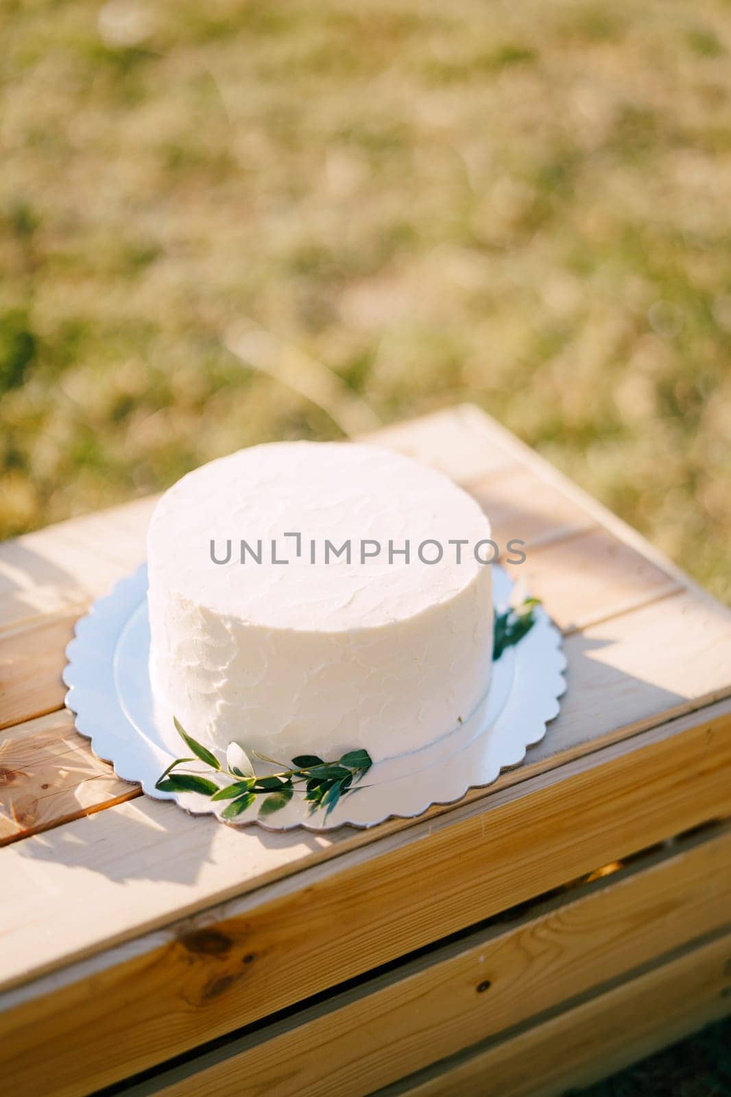 White wedding cake on a plate stands on a wooden box on a green lawn by Nadtochiy