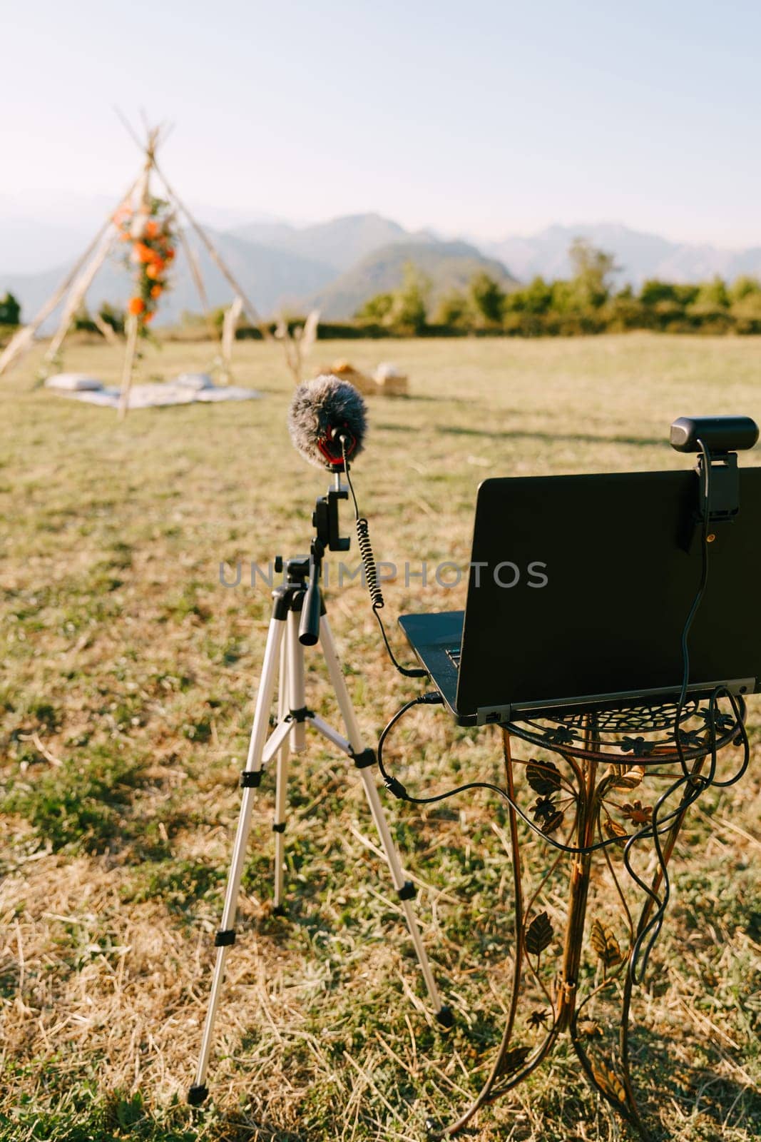 Laptop with a webcam and a microphone on a stand stands in front of a wedding arch on a green meadow. High quality photo