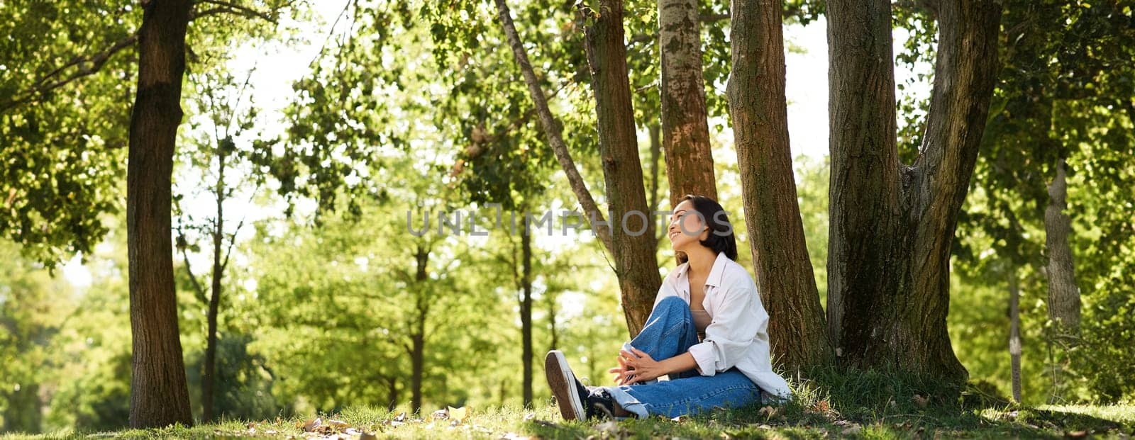 Portrait of asian girl relaxing, leaning on tree and resting in park under shade, smiling and enjoying the walk outdoors.